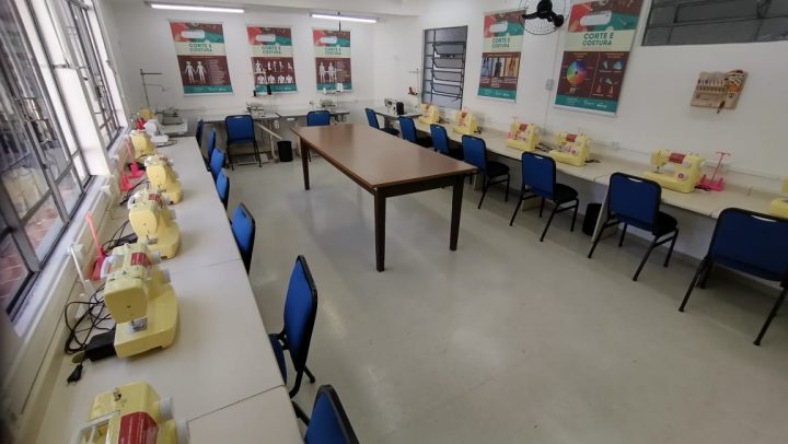 Sala ampla, com 12 máquinas de costura dispostas nas laterais e cadeiras estofadas azuis. Há cartazes sobre corte e costura nas paredes e, ao centro, uma mesa grande usada para criação de moldes e finalização de peças de roupa