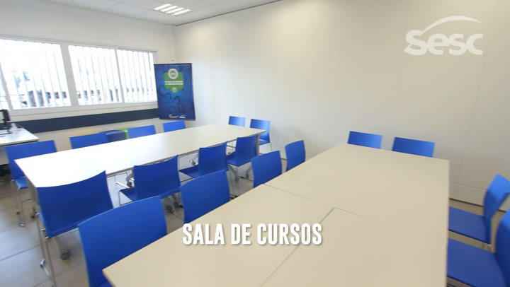 Sala ampla de cursos com 10 mesas e 20 cadeiras azuis. Possui quadro branco, janela grande, computador e armário.